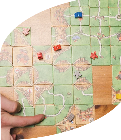 Een hand legt een tegel op het bord van het bordspel Carcassonne, waarop aaneengesloten wegen en velden te zien zijn. Over het bord liggen kleurrijke speelstukken verspreid, wat wijst op strategisch spel.